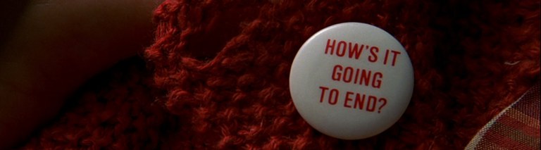 Screenshot from the movie "The Truman Show", showing a zoomed-in-shot of the button badge which states "How's it going to end".